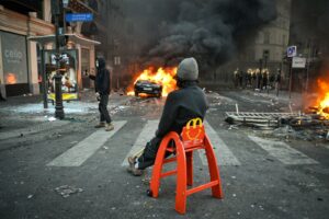 A person sitting on a red high chair in a street with a burning car